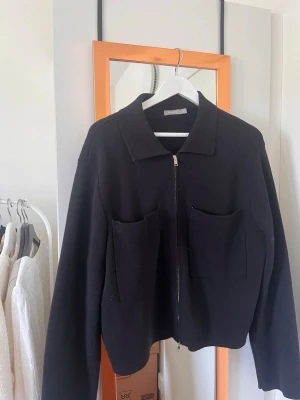 Svart overshirt från Åhléns - Stilren svart overshirt från Åhléns med dragkedja framtill och två stora fickor på bröstet. Jackan har klassisk krage och är långärmad, perfekt för lager på lager. Materialet känns mjukt och har en ullig känsla.