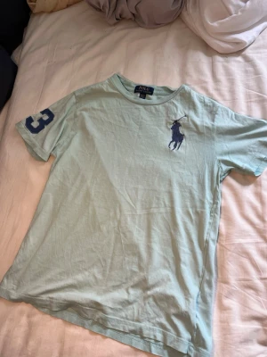 Ljusblå Polo Ralph Lauren t-shirt - Snygg ljusblå t-shirt från Polo Ralph Lauren med stort marinblått logotryck på bröstet och siffran 3 på ärmen. Klassisk passform och rund halsringning, perfekt till jeans eller shorts. 