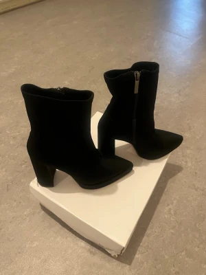 Aldo, Svarta stövletter med klack - Snygga svarta stövletter från Aldo med spetsig tå och kraftig klack. Dragkedja på sidan för enkel påtagning. Perfekta för dig som vill ha en stilren och trendig look. Materialet ser ut att vara mocka eller liknande tyg.