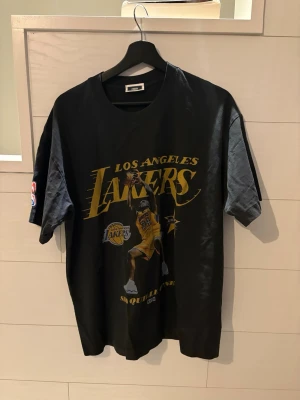 Svart Lakers t-shirt med tryck Kith - Cool svart t-shirt med Los Angeles Lakers-tryck och stor basketspelare på framsidan. Klassisk passform med korta ärmar och rund hals. Perfekt för dig som gillar basket och streetwear.