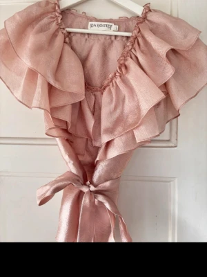 Rosa volangblus från Ida Sjöstedt - Superfin rosa blus från Ida Sjöstedt med stora volanger och knytband i midjan. Blusen har en glansig finish och romantisk känsla, perfekt för dig som gillar statement-plagg. Materialet är lätt och skimrande, vilket ger en lyxig look.