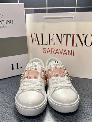 Valentino Garavani vita sneakers med nitar - Snygga vita sneakers från Valentino Garavani med breda ljusrosa band och coola pyramidformade nitar på sidorna och hälen. Skorna har en chunky sula och klassisk snörning. Perfekta för dig som vill sticka ut med en lyxig och edgy stil.