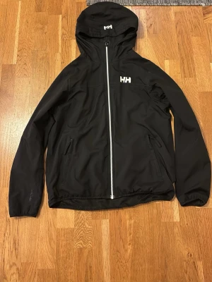 Svart vindjacka från Helly Hansen - Snygg svart vindjacka från Helly Hansen med vit dragkedja och logga på bröstet och huvan. Jackan har huva, två sidofickor med dragkedja och justerbar nederkant. Lite använd men i bra skick utan skråmor eller smuts. Är nog storlek S då den passar mig bra som 175 cm lång