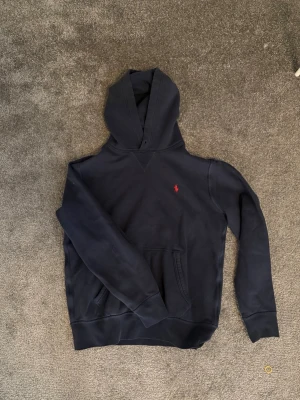 Polo Ralph Lauren Hoodie - Snygg mörkblå hoodie från Polo Ralph Lauren med klassisk röd logga broderad på bröstet. Hoodien har huva, känguruficka och ribbade muddar vid ärmar och nederkant. Perfekt för en chill och stilren look. Snörerna borttagna men inget man tänker på vid användning. Storlek S 