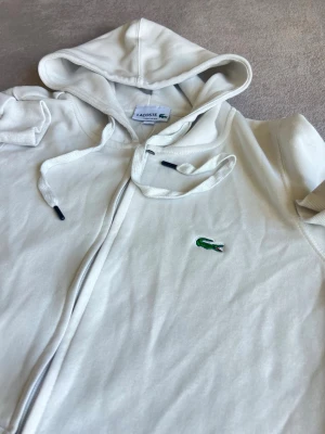 Vit hoodie från Lacoste med dragkedja - Säljer en vit hoodie från Lacoste med dragkedja framtill och klassisk krokodillogga på bröstet. Hoodien har huva med dragsko och två fickor framtill. Tillverkad i mjukt bomullsmaterial, perfekt för en avslappnad stil.