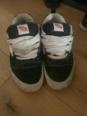 Svarta och vita Vans sneakers - Klassiska Vans sneakers i svart och vitt med breda vita skosnören och tydlig Vans-logga på plösen. Skorna har låg profil och vit sula, samt kontrastsömmar som ger en snygg streetwear-look. Ovandelen är i mocka och textil.
