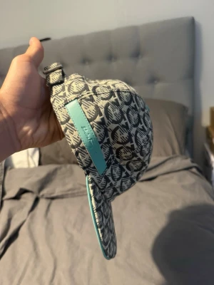 Ferragamo keps - Säljer en snygg keps från Salvatore Ferragamo med grått och svart mönster samt turkosa detaljer och logga på sidan. Kepsen har böjd skärm och justerbart spänne bak. Materialet känns som bomullscanvas och detaljerna är i läder.
