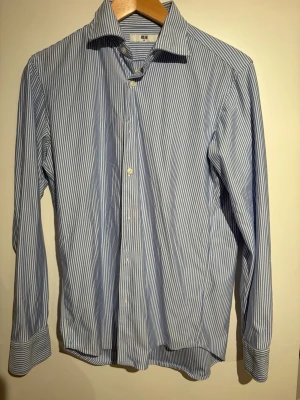 Blåvit randig skjorta från Uniqlo - Klassisk blå och vit randig skjorta från Uniqlo i storlek XS. Skjortan har lång ärm, knappar framtill och en stilren krage. Perfekt för en clean och fräsch look. Tillverkad i mjuk bomull för skön komfort.