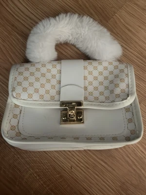 Vit handväska med fluffigt handtag - Säljer en vit handväska med guldigt lås och ett fluffigt vitt handtag. Väskan har ett mönster med små cirklar och detaljer i beige. Materialet ser ut att vara konstläder och handtaget är i fuskpäls. Perfekt för dig som vill ha en unik accessoar!