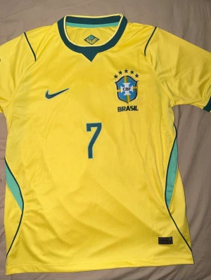 Brasilien fotbollströja Vini Jr #7 VM 2026 - Gul Brasilien fotbollströja med nummer 7 och Vini Jr. på ryggen. Tröjan har gröna och blå detaljer, korta ärmar och officiella FIFA World Cup 2026-märket på ärmen. Tillverkad i lätt och ventilerande polyester, perfekt för fotbollsfans.