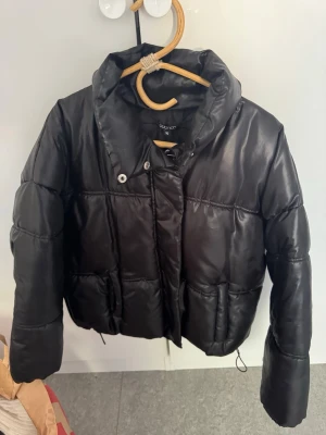 Svart pufferjacka från Boohoo - Snygg svart pufferjacka från Boohoo med hög krage och stora fickor framtill. Jackan har en glansig finish och stängs med dragkedja och tryckknappar. Perfekt för kalla vinterdagar och ger en trendig look.