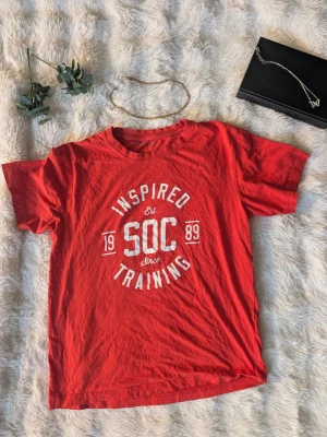 Röd t-shirt med tryck från SOC - Röd t-shirt från SOC med vitt tryck framtill där det står 'Inspired Training Est 1989'. Klassisk rund hals och korta ärmar. Perfekt för dig som gillar sportig och avslappnad stil.