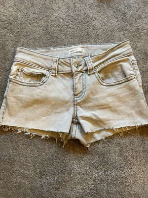 Ljusgrå jeansshorts med rå kant - Säljer ett par ljusgrå jeansshorts med rå, fransig kant och klassisk femficksdesign. Shortsen har knappgylf, bälteshällor och bakfickor med lock och knapp. Perfekta för varma dagar och enkel att matcha med allt.