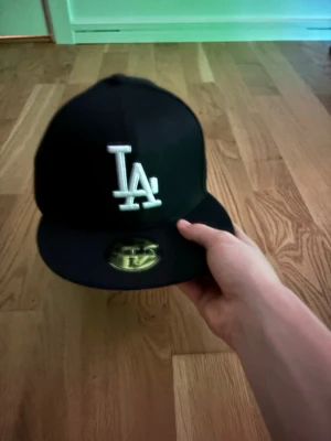 Svart LA-keps från New Era - Säljer en svart fitted keps från New Era med LA-logga broderad i vitt framtill. Klassisk 59FIFTY-modell med platt skärm och guldigt klistermärke på ovansidan. Snygg streetwear-vibe och passar perfekt till en avslappnad stil.