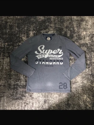 Grå långärmad tröja från Superdry - Snygg grå långärmad tröja från Superdry med tryck i vitt och mörkgrått framtill. Rund halsringning och mjukt material som passar perfekt till jeans. Stort tryck med texten 'Super Dry Japan Registered Standard Issue' och siffran 28 nere till vänster. Passformen är lite mer relaxed. Pris kan såklart diskuteras!
