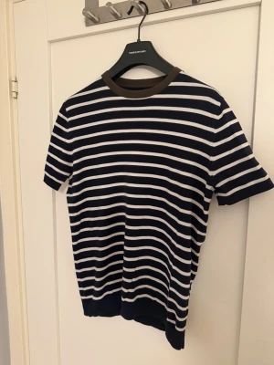 Randig t-shirt från Massimo Dutti - Snygg randig t-shirt från Massimo Dutti i marinblått och vitt. T-shirten har korta ärmar, rund halsringning med kontrasterande brun kant och är tillverkad i mjuk bomull. Perfekt för en avslappnad och stilren look.