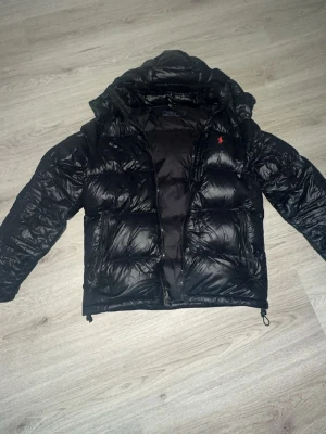 Svart dunjacka från Polo Ralph Lauren - Snygg svart dunjacka från Polo Ralph Lauren med röd broderad logga på bröstet och RL-dragkedja. Jackan har huva, två sidofickor och en puffig, quiltad design som håller dig varm under kalla dagar. En liten defekt kom dm för bild