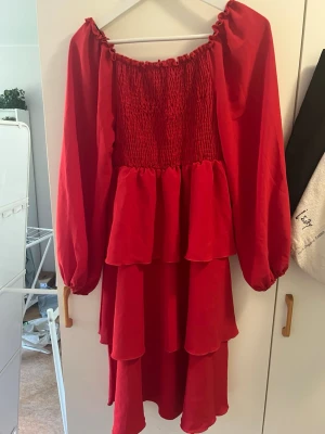 Röd volangklänning med långa ärmar - Snygg röd klänning med volanger och långa puffärmar. Klänningen har smock upptill och en offshoulder-design som ger en trendig look. Perfekt för dig som vill sticka ut med färg och stil.