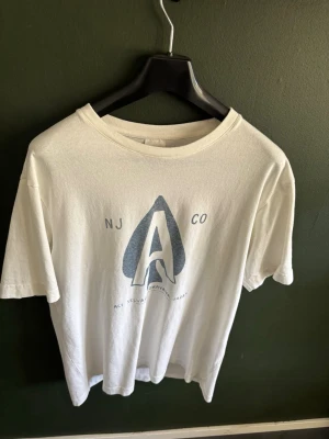 Nudie t shirt - Vit t-shirt med ett stort tryck framtill i blått med bokstaven A och texten NJ CO. Klassisk rund hals och korta ärmar. Tillverkad i mjuk bomull för en skön känsla. Perfekt till jeans eller shorts för en avslappnad look.