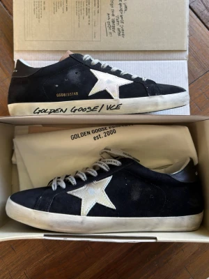 Golden Goose svarta sneakers  - Snygga svarta sneakers från Golden Goose med silver stjärna på sidan och vit sula. Skorna har grå skosnören och detaljer i svart läder på hälen där märket står tryckt. Klassisk låg modell med slitna detaljer för en najs look.