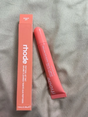Rhode Peptide Lip Tint Peach Pit - Peptide Lip Tint från Rhode i färgen Peach Pit. Läppbalsamet kommer i en mjuk tub med skruvkork och har en persikofärgad ton. Förpackningen är stilren och modern med vit text. Perfekt för att ge läpparna en fräsch och återfuktad look.