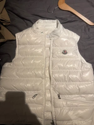 Vit dunväst från Moncler - Snygg vit dunväst från Moncler med glansig finish och klassisk logga på bröstet. Västen har hög krage, dragkedja och tryckknapp framtill samt två fickor med dragkedja. Perfekt för lager på lager och streetstyle.