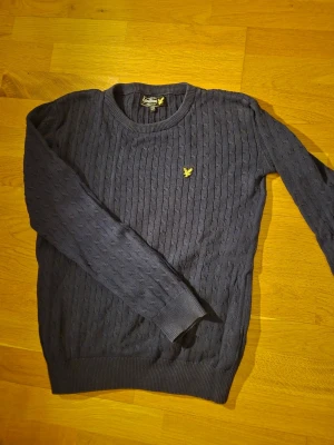Mörkblå kabelstickad tröja Lyle & Scott - Mörkblå kabelstickad tröja från Lyle & Scott med rund halsringning och gul logga broderad på bröstet. Tröjan har ribbade muddar vid ärmslut och nederkant. Perfekt för dig som gillar klassisk och stilren design.