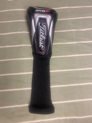 Titleist 915F headcover svart/vit - Snygg headcover till golfklubba från Titleist, modell 915F. Färgerna är svart, vit och lite rött. Ovandelen är i konstläder med broderad logga och modellen står tydligt. Nedre delen är i stretchigt stickat material för enkel på- och avtagning.