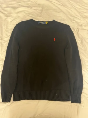 Svart stickad tröja från Polo Ralph Lauren - Klassisk svart stickad tröja från Polo Ralph Lauren med rund halsringning och den ikoniska röda logotypen broderad på bröstet. Tröjan har långa ärmar och ribbade muddar vid ärmslut och nederkant. Perfekt till höst och vinter.