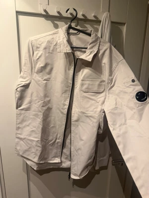 Cp company  - Snygg vit vindjacka med dragkedja framtill, bröstficka och en detalj på ärmen. Jackan har en klassisk krage och raka ärmar. Materialet ser ut att vara lätt och vindtätt, perfekt för blåsiga dagar.