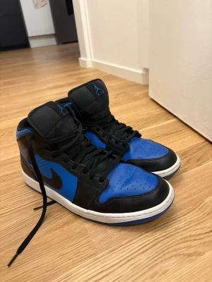 Svart/blåa Air Jordan 1 sneakers - Säljer ett par Air Jordan 1 sneakers i svart och blått med vita sulor. Skorna har högt skaft, klassisk Jordan-logga på plösen och sidopanelen samt svarta skosnören. Materialet är en mix av läder och syntet. Perfekta för dig som gillar streetwear och ikoniska sneakers.