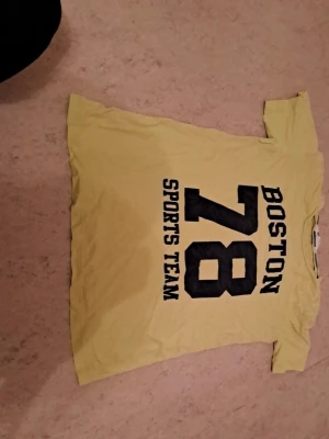 Gul Boston 78 t-shirt - Gul t-shirt med svart tryck på ryggen där det står 'BOSTON 78 SPORTS TEAM' i stora bokstäver. Klassisk passform och korta ärmar, perfekt för dig som gillar sportig stil. Tillverkad i mjukt bomullstyg för skön känsla hela dagen. I bra skick. Säljs för 20 kr. Tveka inte att kontakta om du är intresserad eller har några frågor! Först till kvarn dom gäller!