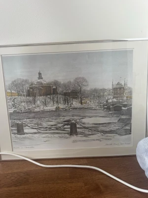 Inramad vintertavla Stockholm - Säljer en inramad tavla med vintermotiv från Stockholm, föreställande en snötäckt stadsmiljö med kyrka och bro. Trycket är signerat av konstnären och har en ljus passepartout samt vit ram. Perfekt för dig som gillar konst och stadsmotiv.