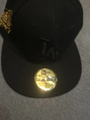 Svart LA 59FIFTY keps - Säljer en svart New Era 59FIFTY keps med LA-logga framtill och gul detalj på sidan. Klassisk flat brim och broderade ventilationshål. Kepsen är i fast modell och har en guldfärgad sticker på skärmen.
