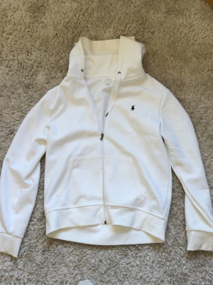 Vit hoodie från Polo Ralph Lauren - Snygg vit hoodie med dragkedja från Polo Ralph Lauren. Klassisk design med svart broderad logga på bröstet, huva med dragsko och känguruficka framtill. Tillverkad i mjukt bomullsmaterial och har ribbade muddar vid ärmar och nederkant.