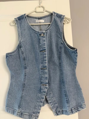 Ärmlös jeansväst i ljus denim - Snygg ärmlös jeansväst i ljusblå denim med knappar framtill och figurnära passform. Västen har rund halsringning och markerade sömmar som ger en cool vintagekänsla. Perfekt att styla över en t-shirt eller topp för en trendig look.