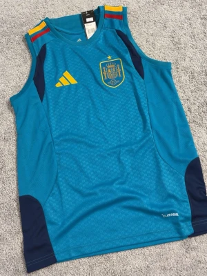 Blå Spanien linne från Adidas - Säljer ett blått sportlinne från Adidas med Spaniens landslagsemblem i gult på bröstet. Linnet har gula och röda detaljer på axlarna, samt mörkblå partier vid sidorna. Tillverkat i lätt och ventilerande ClimaCool-material, perfekt för träning eller match.
