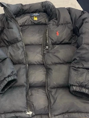 Svart pufferjacka från Polo Ralph Lauren - Kom med frågor priset kan diskuteras Snygg svart pufferjacka från Polo Ralph Lauren med röd broderad logga på bröstet. Jackan har hög krage, dragkedja framtill och elastiska muddar vid ärmsluten. Perfekt för kalla vinterdagar och har en klassisk, boxig passform.