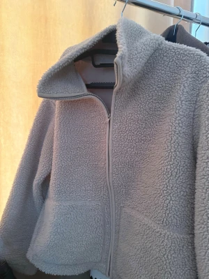Beige teddyjacka med dragkedja - Mysig beige teddyjacka med hög krage och hel dragkedja framtill. Jackan har två stora fickor och är gjord i fluffigt, mjukt material som ger en avslappnad och trendig look. Perfekt för kyliga dagar när du vill vara både varm och stylish.