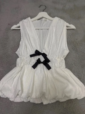 Vit topp med svarta rosetter - Söt vit topp med v-ringad hals och två svarta rosetter framtill. Västen har en volangliknande nederdel och är tillverkad i ett strukturerat tyg som ger en unik look. Perfekt för att lyfta en outfit med en lekfull touch.