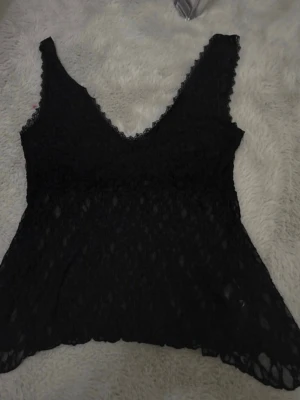Svart spetslinne med v-ringning - Säljer ett svart linne i spets med djup v-ringning både fram och bak. Toppen är ärmlös och har ett transparent, mönstrat spetsmaterial som ger en snygg och trendig look. Perfekt att styla lager på lager eller till ett par jeans.