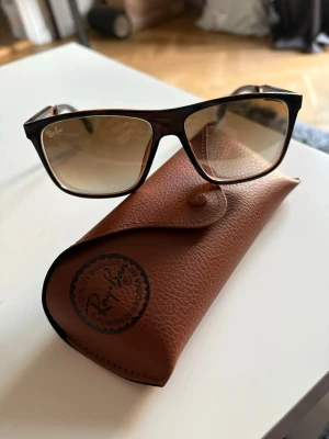 Bruna Ray-Ban solglasögon i retrostil - Säljer ett par snygga Ray-Ban solglasögon med bruna bågar och tonade glas. Modellen har en klassisk retroform och kommer med ett brunt fodral i skinnimitation. Perfekta för soliga dagar och ger en stilren look.