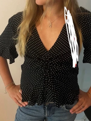 Svart prickig blus med snörning - Snygg svart blus med vita prickar och v-ringning. Toppen har snörning framtill som ger en cool rynkad effekt och volangiga ärmar i trekvartslängd. Perfekt till jeans eller kjol för en trendig look.💗