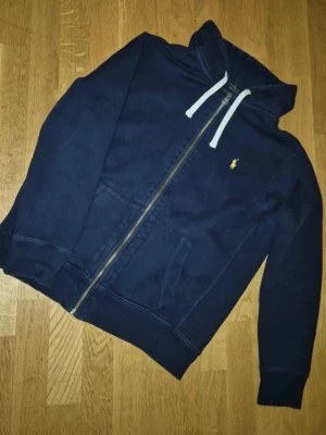 Hoodie med dragkedja Polo Ralf Lauren - Snygg mörkblå hoodie från Polo Ralph Lauren med dragkedja framtill, vita dragsnören i huvan och klassisk gul logga på bröstet. Hoodien har känguruficka och ribbade muddar. Perfekt för en avslappnad och stilren look.