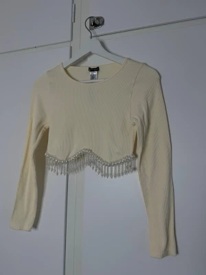 Vit croppad topp med pärlor - Trendig croppad topp i ribbad vit bomull med långa ärmar. Toppen har en unik vågig kant nedtill dekorerad med pärlor som ger en extra touch. Perfekt för dig som vill sticka ut med detaljer.