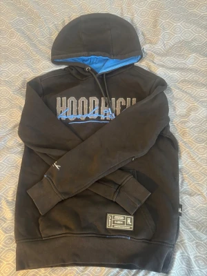 Svart hoodie från Hoodrich med blå detaljer - Säljer en svart hoodie från Hoodrich med stor logga i vitt och blått på bröstet och ryggen. Insidan av huvan är blå och det finns en praktisk magficka framtill. Stora muddar vid ärmslut och nederkant samt snygga detaljer och patch nertill.
