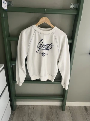 Vit sweatshirt New York Giants NFL - Vit sweatshirt med blå text och tryck från New York Giants och NFL-logga på bröstet. Tröjan har rund halsringning, långa ärmar och ribbade muddar. Perfekt för dig som gillar sportig stil och amerikansk fotboll.