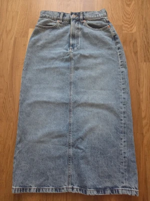 Lång jeanskjol i ljusblå denim - Snygg lång jeanskjol i ljusblå denim med klassiska femficksdetaljer och hög midja. Kjolen har rak passform och slits bak för extra rörelsefrihet. Perfekt för en avslappnad och trendig look.