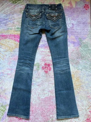 Miss me lågmidjade utsvängda jeans - Snygga vintage blåa bootcut jeans med låg midja från MissMe😍De har slitningar och dekorerade bakfickor med nitar, diamanter, pärlor ich broderier! Klassisk femficksmodell med bakfickor som kan stängas med knapp🩷Perfekta för dig som vill ha en cool & unik look. Passar bra till alla plagg, vardagen eller finare events🙌Så vackra som verkligen får din outfit på topp! Storlek W24/L34, passar XS då de är stretchiga.  Mått cm: midja 34, gren 18, innerben 85💋Älskar de färgglada detaljerna & passformen❤️