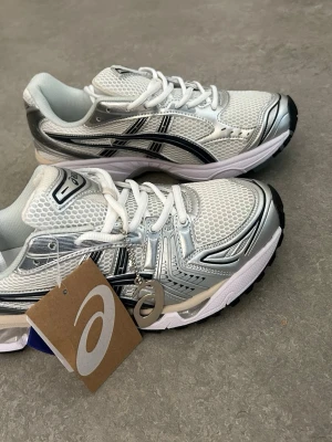 Vita och silver Asics sneakers - Snygga sneakers från Asics i vitt och silver med svarta detaljer. Ovandelen är i mesh och syntetmaterial för bra andningsförmåga och komfort. Klassisk låg modell med vita skosnören och svart yttersula. Perfekta för en sportig och trendig look.